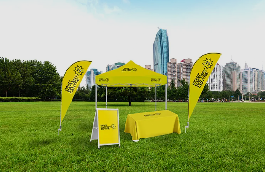 Bright yellow outdoor event booth set up on a grassy field, featuring a branded canopy tent, two matching feather flags, a covered table, and a sidewalk sign, with a city skyline in the background.
