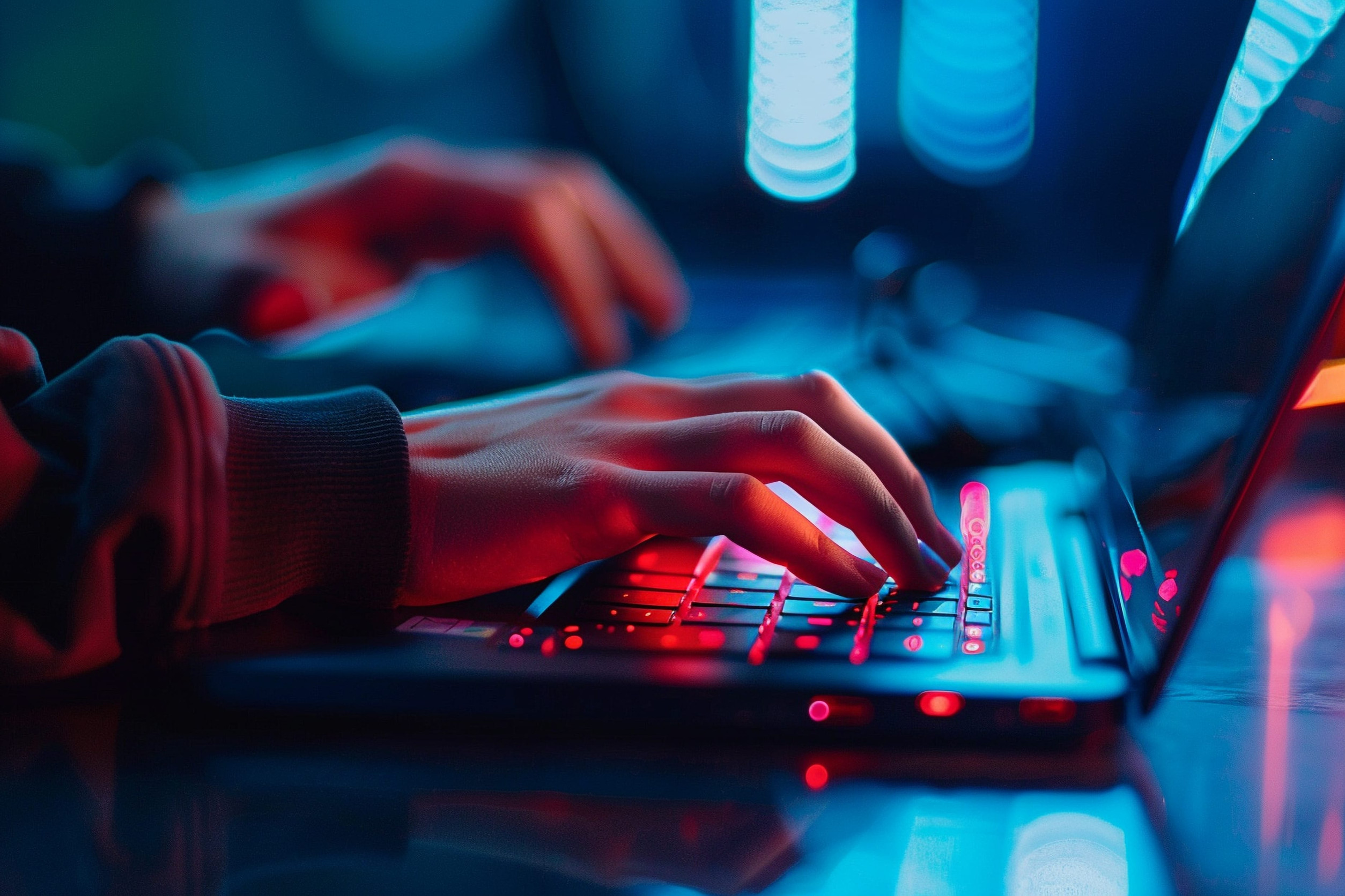 Close-up of hands typing on a laptop keyboard lit by vivid blue and red neon light, creating a dramatic high-tech atmosphere.
