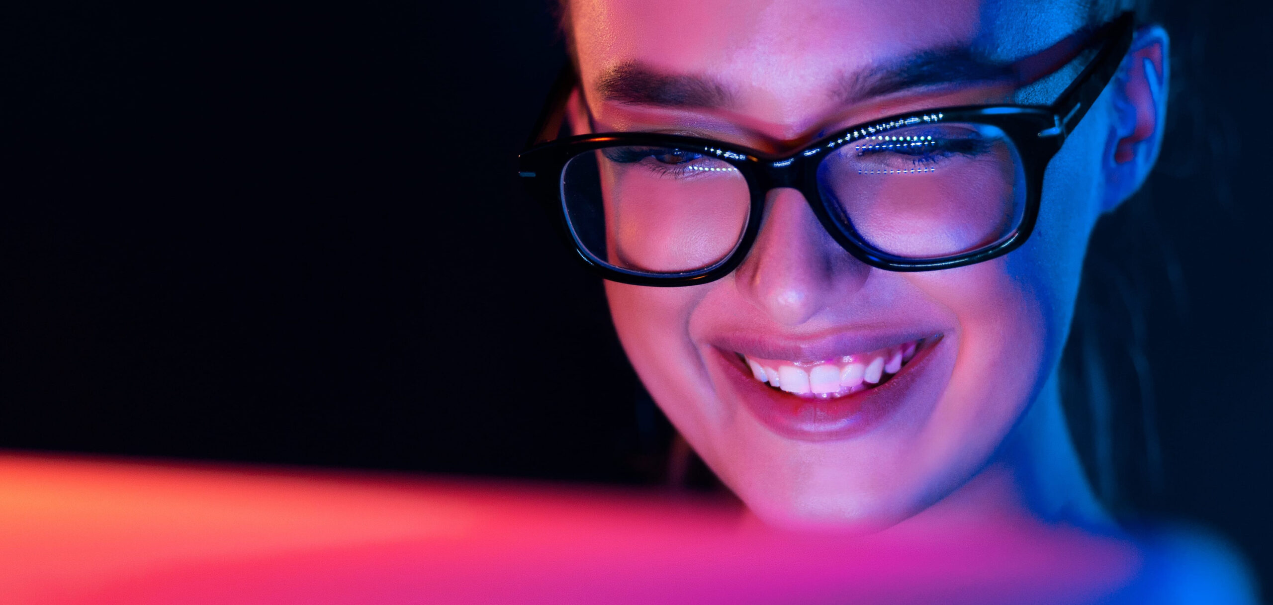 Close-up of a smiling person wearing glasses, illuminated by vivid pink and blue neon light while looking at a screen in a dark setting.