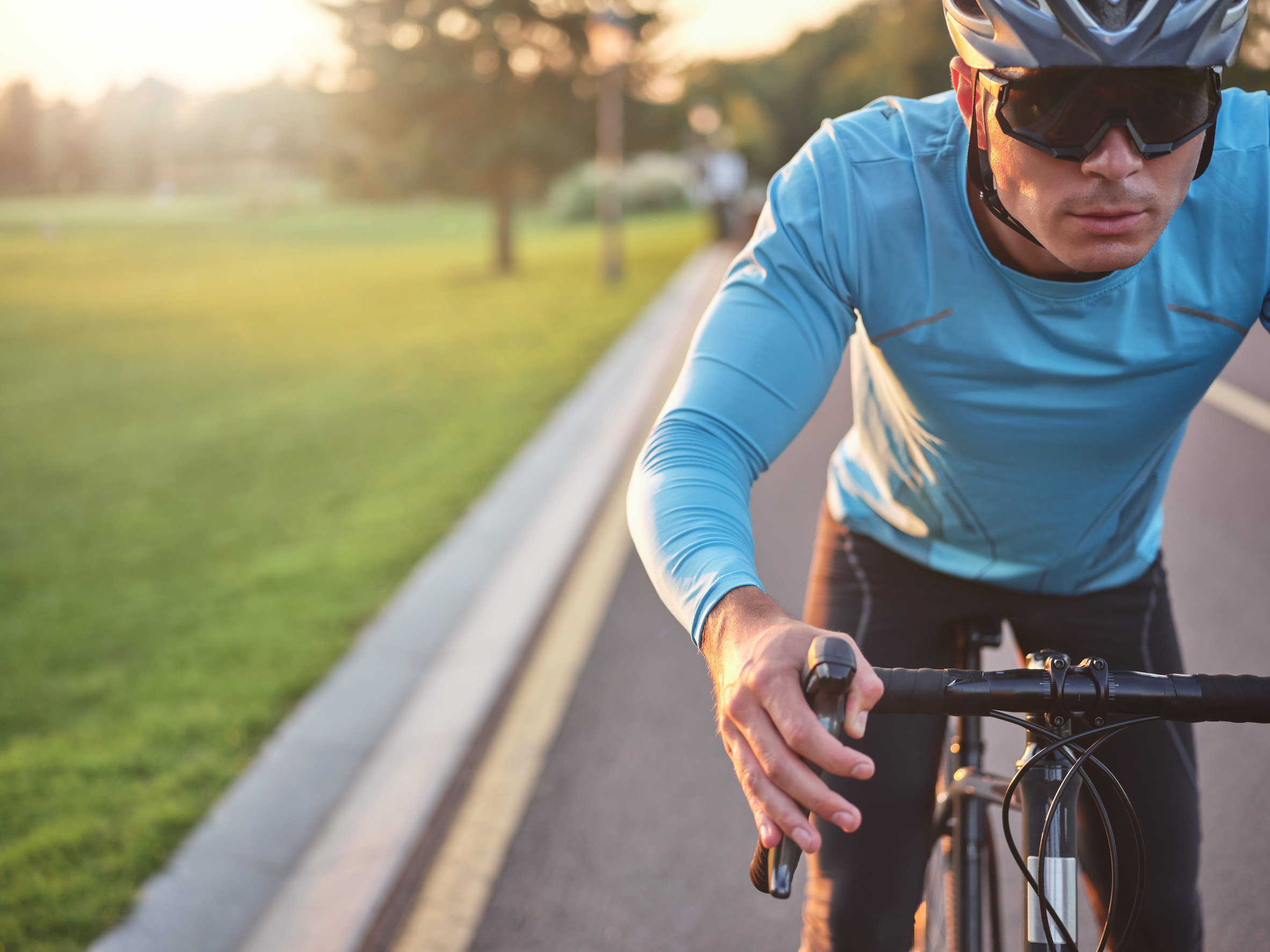 Close-up of a cyclist in a blue long-sleeve jersey, helmet, and sunglasses riding on a road at sunset, with one hand on the handlebar and a grassy park blurred in the background.