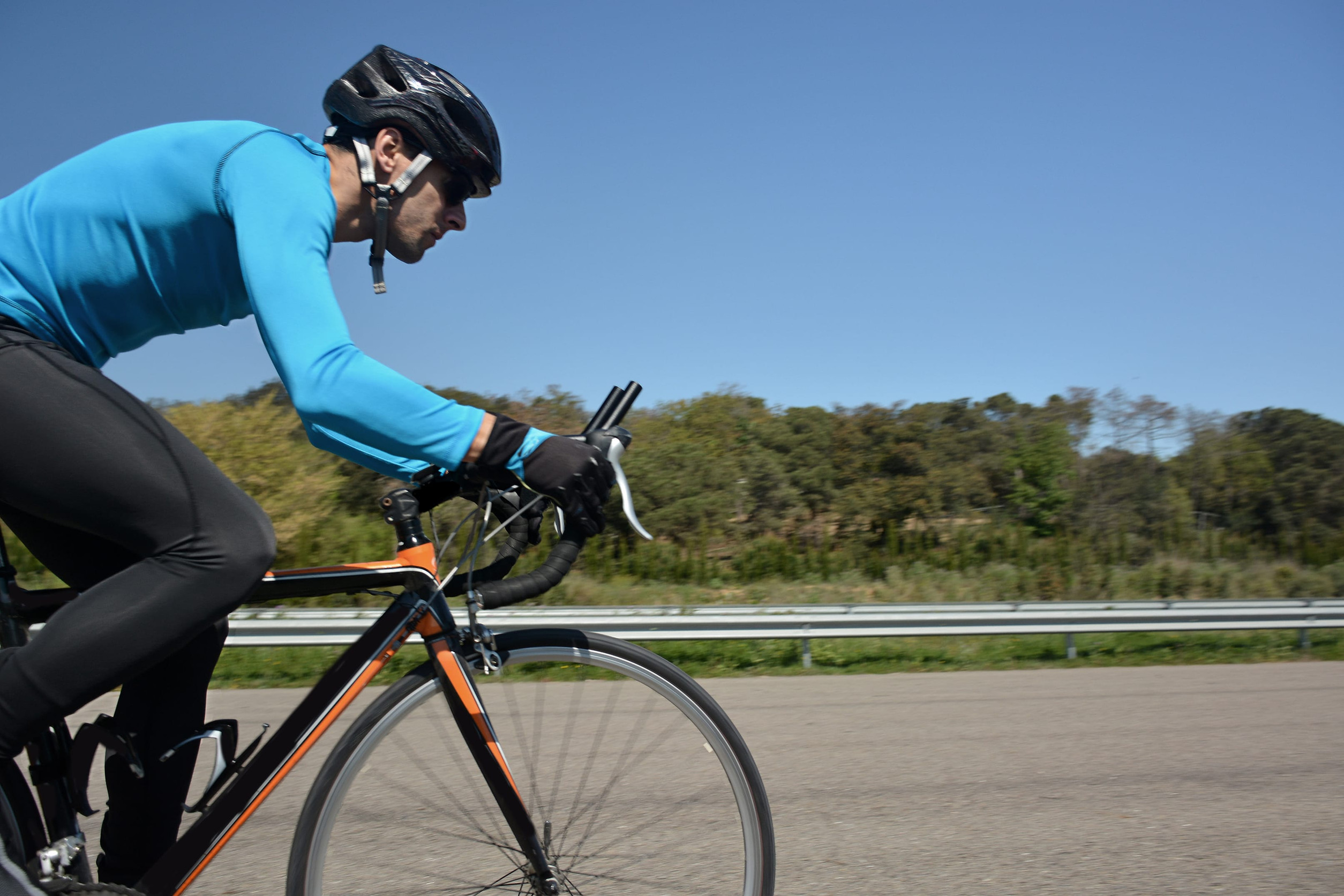 Close-up side view of a cyclist in a blue long-sleeve jersey and black helmet riding a road bike on a paved road, with trees and a clear blue sky in the background.