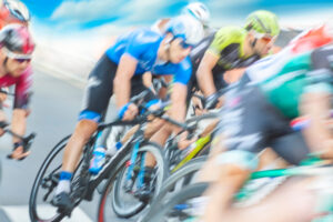 Motion-blurred group of road cyclists racing closely together on a paved course, wearing colorful helmets and jerseys that emphasize speed and intensity.