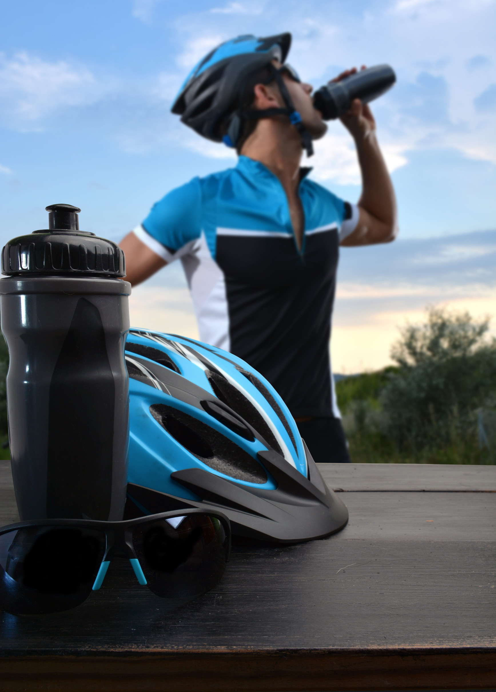 Blue cycling helmet, water bottle, and sunglasses on a table in the foreground, with a cyclist in a matching blue and black jersey drinking from a bottle outdoors at sunset.