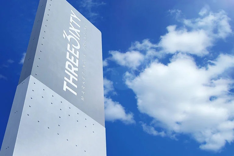 Tall rectangular outdoor sign with the THREE6IXTY Marketing & Branding logo against a bright blue sky with scattered white clouds.