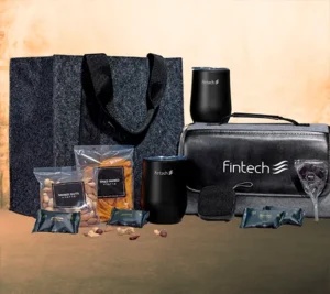 Branded corporate gift set arranged on a table, including a textured tote bag, two black tumblers, a black zip pouch with the “Fintech” logo, and packaged snack items.