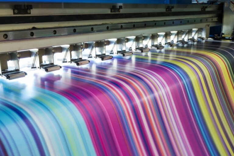 **Alt text:** Large-format industrial printer producing vibrant multicolored print material, with rows of print heads applying bright streaks of ink across a moving surface.